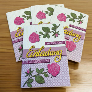 Vier Karten zum 70. Geburtstag mit rosa Rosen und gelben handgeschriebenem Titel "Einladung" auf einem Holztisch in einem Blogbeitrag über Einladungen.