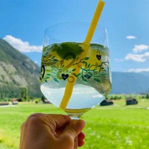 Ein Bild von einem Glas mit gelbem Strohhalm und im Hintergrund sieht man die grünen Wiesen und den blauen Himmel des Tiroler Oberlands in einem Blogbeitrag über den Sommer in Tirol im Juli.
