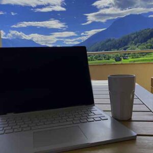 Ein Laptop und ein Kaffeebecher stehen auf einem Balkon mit Aussicht auf einen blauen Himmel.