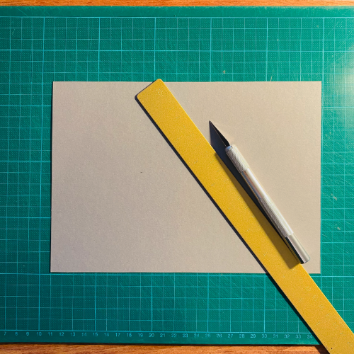 Ein grauer Bastelkarton mit einem goldenen Lineal und einem Cutter auf einer grünen Schneidunterlage in einem Blogbeitrag über selbstgebastelte Babykarten.