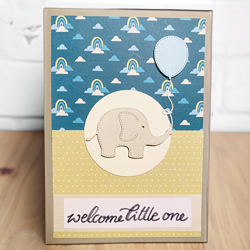 Eine Grußkarte im Scrapbooking-Stil mit Regenbogen-Musterpapier, einem Elefanten und einem Ballon aus Papier und dem hangeschriebenen Schriftzug "welcome little one" in einem Blogbeitrag über selbstgebastelte Babykarten.
