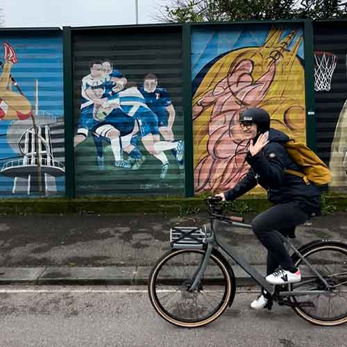 Eine Person auf einem Fahrrad, im Hintergrund sind bunte Graffiti zu sehen. Die Person winkt und hat einen gelben Rucksack auf dem Rücken.