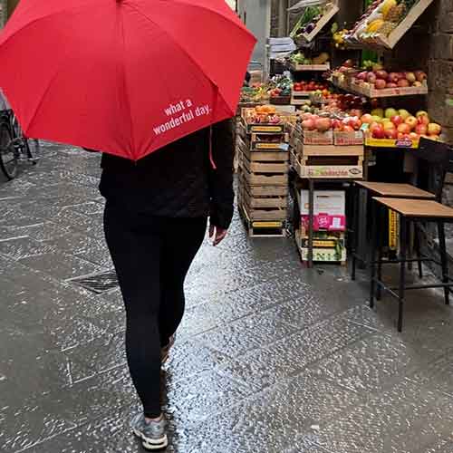 Eine regennasse Gasse mit einer Person, die einen roten Schirm aufgespannt hat. Neben ihr sind bunte Obstkisten zu sehen.