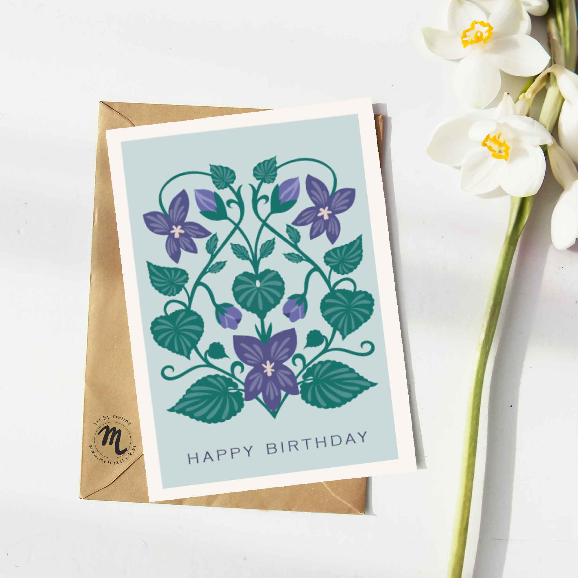 Mock-up einer Grußkarte mit einer Illustration von Veilchen und happy birthday-Schriftzug. Daneben liegt eine weiße Blume.