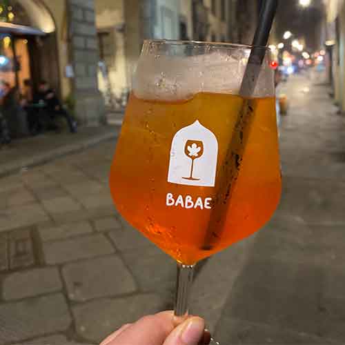 Ein Aperol Sprizz mit einem schwarzen Strohhalm wird in der Hand gehalten, dahinter ist eine Straße bei Nacht zu sehen.