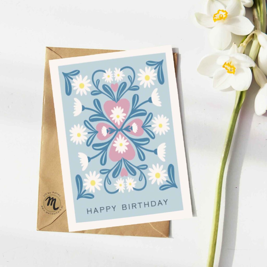 Eine Grußkarte mit "happy birthday"-Schriftzug und illustrierten Gänseblümchen im Folkart-Stil liegt auf einem braunen Kuvert. Daneben liegt eine weiße Blume.